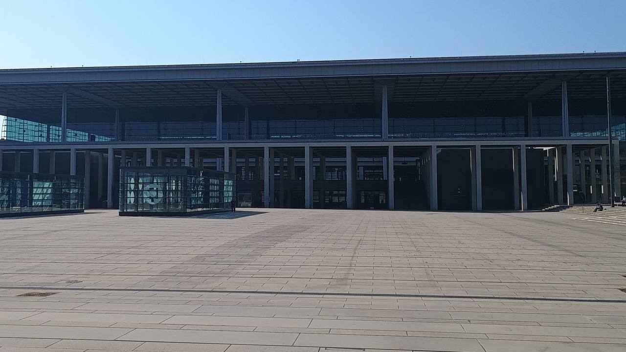 Terminal 1 des Flughafens BER: lange, flache Glasfassade unter einem weit auskragenden Dach, davor der weite, gepflasterte Willy-Brandt-Platz bei blauem Himmel.